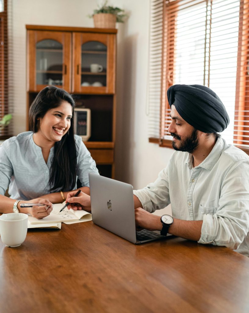 pexels-photo-4308094-4308094 Smiling young Indian man in shirt and turban using laptop at wooden table and helping cheerful female friend while working remotely at home